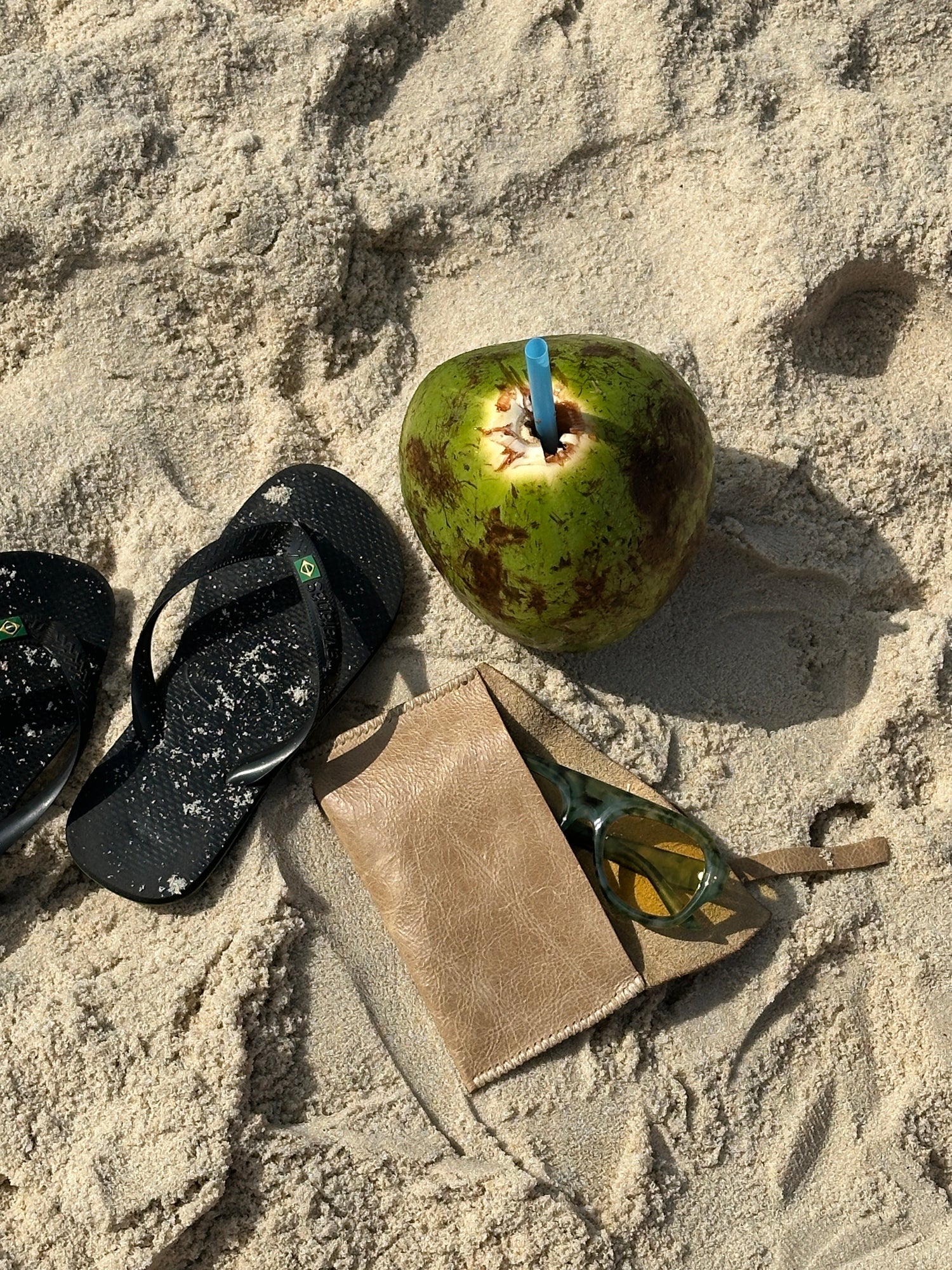 A styled beach scene on sand featuring a green coconut with a blue straw, black flip-flops, a tan leather pouch, and sunglasses.