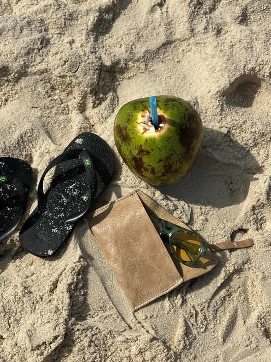 A styled beach scene on sand featuring a green coconut with a blue straw, black flip-flops, a tan leather pouch, and sunglasses.