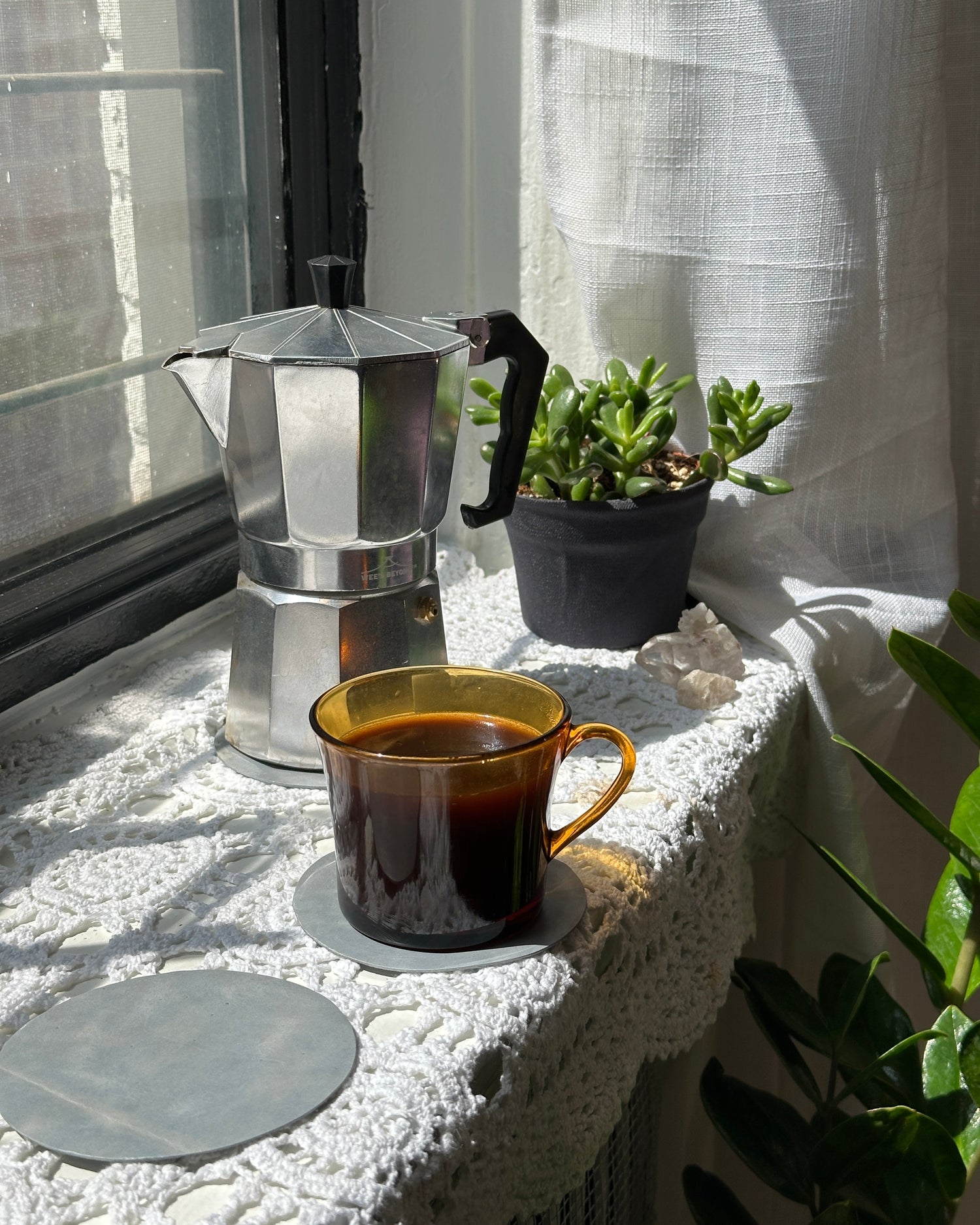 Silver coffee moka pot and yellow tea cup on a white table cloth and light blue leather coasters with plants in the background 