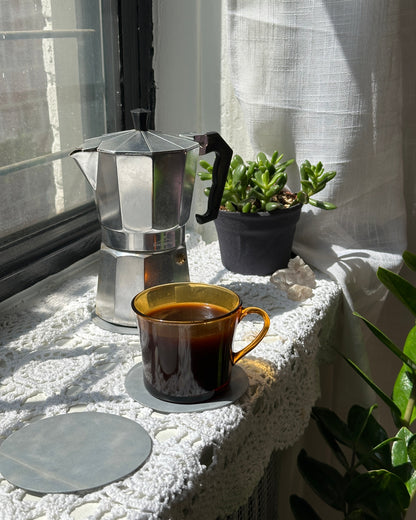 Silver coffee moka pot and yellow tea cup on a white table cloth and light blue leather coasters with plants in the background 
