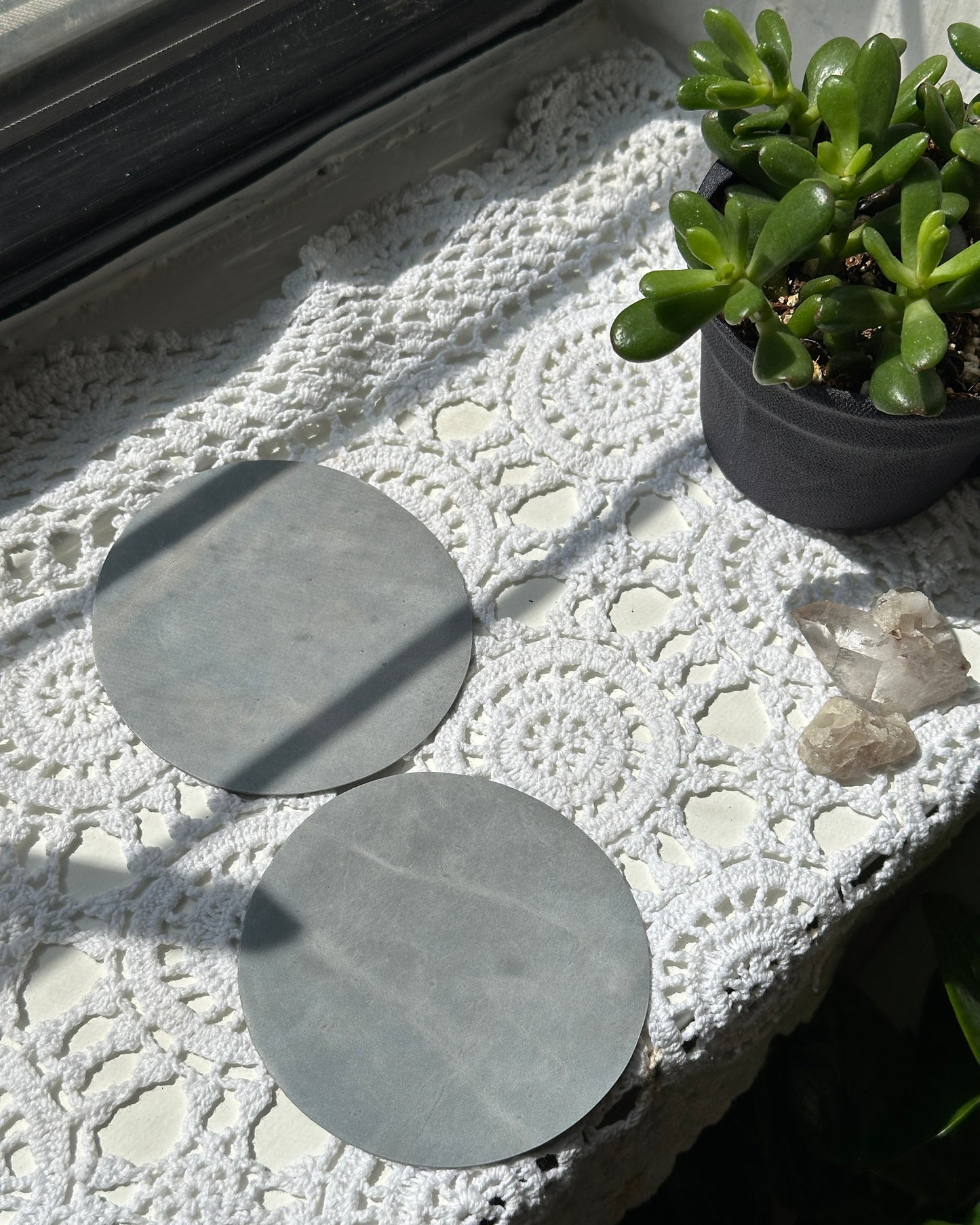 Two light blue leather coasters on a textured surface with plants and crystals in the background