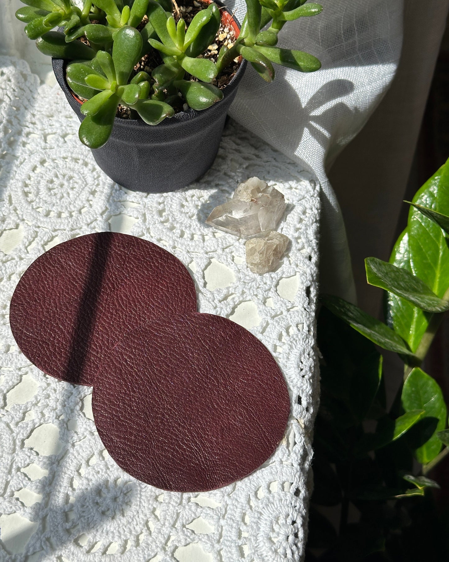 Two burgundy coasters on a textured surface with plants and crystals in the background