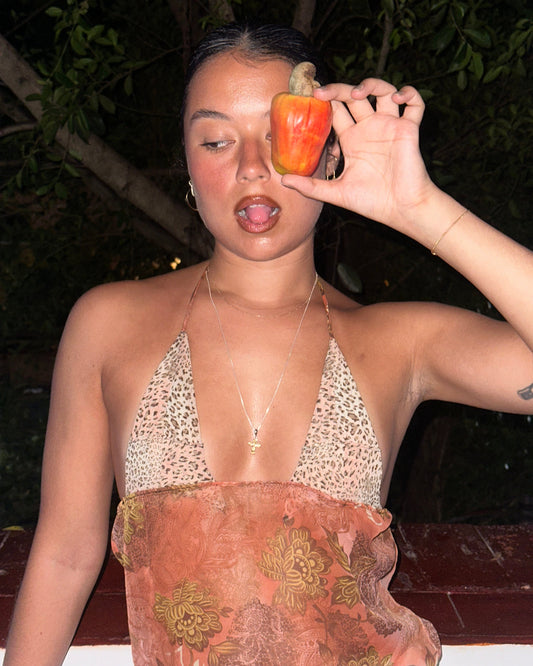 Person holding a caju fruit near their face in a windowsill wearing a caju.nyc tank top inspired by roberto cavalli