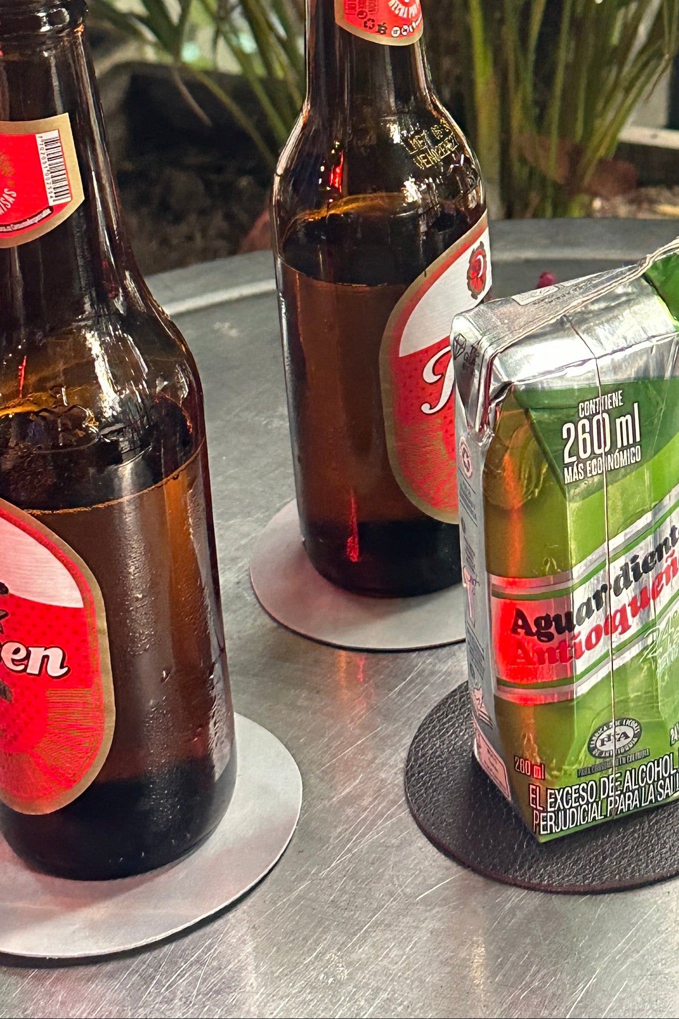 Three brown glass beer bottles with red labels and a green carton of aguardiente resting on a metal tabletop next to plants