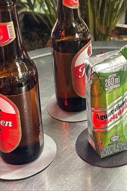 Three brown glass beer bottles with red labels and a green carton of aguardiente resting on a metal tabletop next to plants